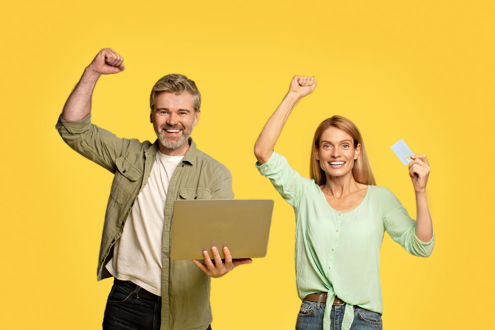 A cheerful man and woman celebrating success with raised fists, holding a laptop and a credit card, against a bright yellow background at home.